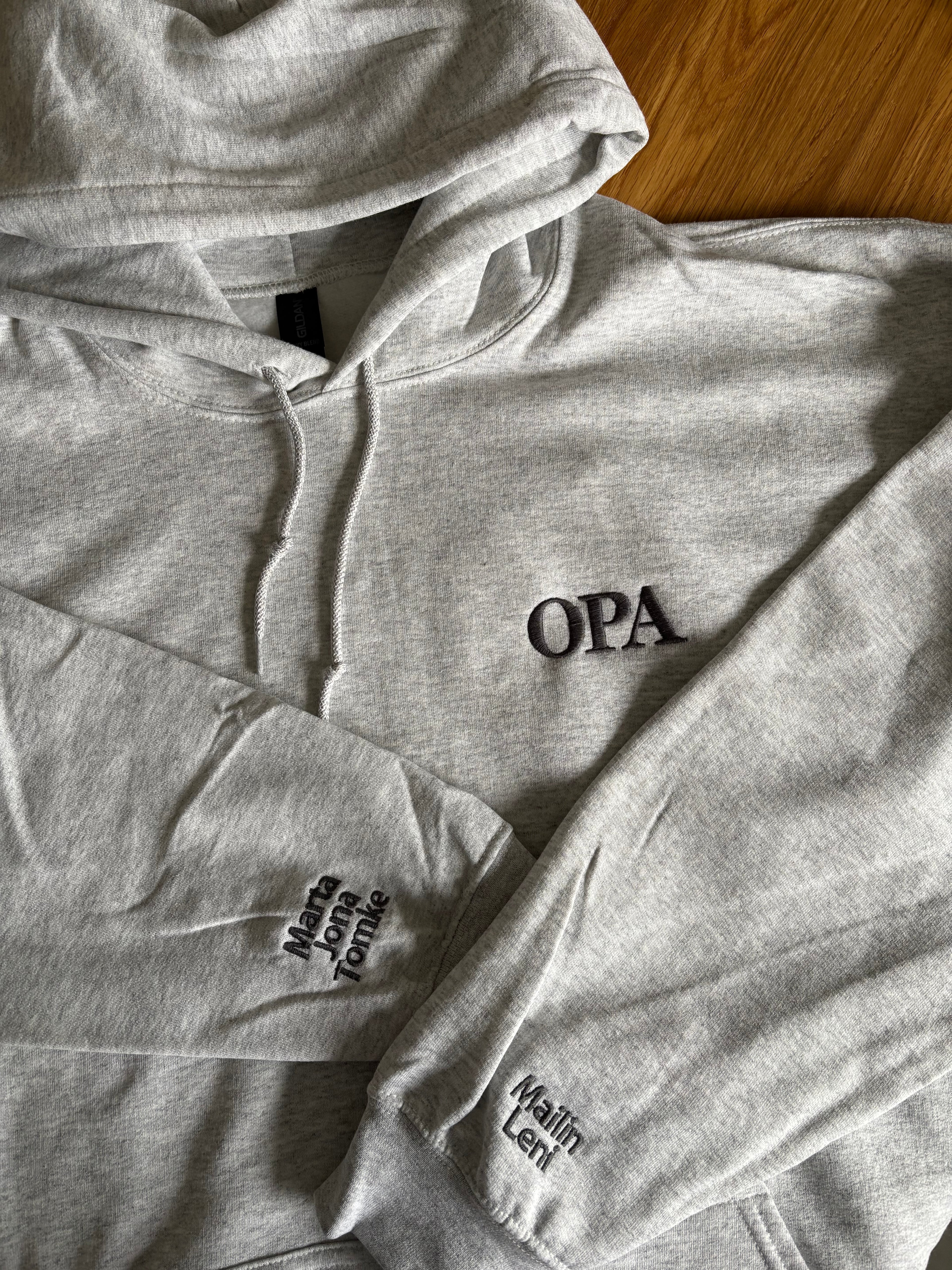 Ein hellgrauer Hoodie mit einem dunkelgrauen Stick "Opa" mit den jeweiligen Kindernamen auf den Ärmeln.