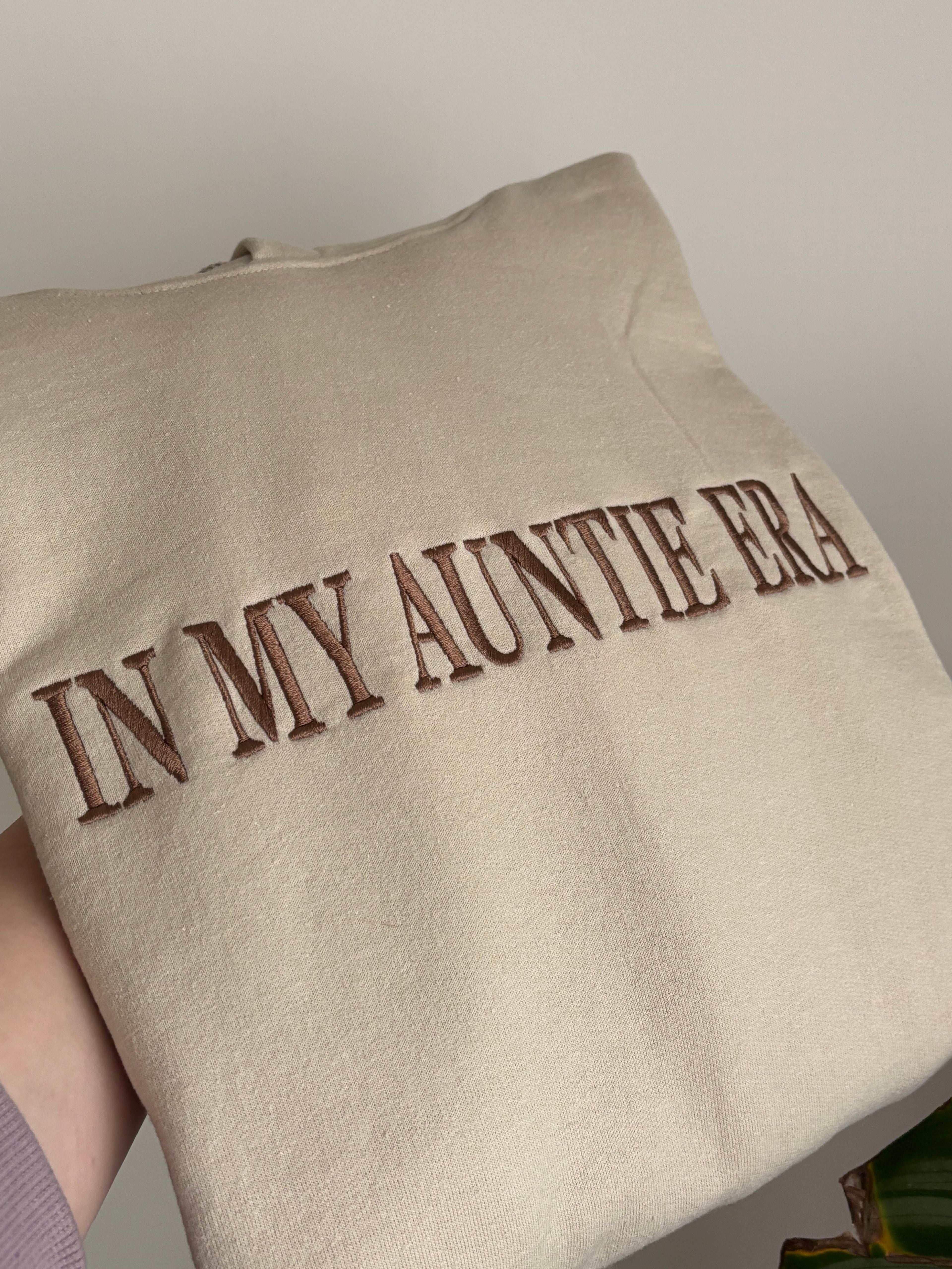 Ein Hoodie in der Farbe Beige mit dem Wunschtext „IN MY AUNTIE ERA“ in der Garnfarbe Brown.