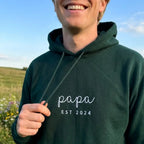 Ein Mann trägt einen personalisierten Hoodie mit einer Stickerei vorne: papa est 2024.