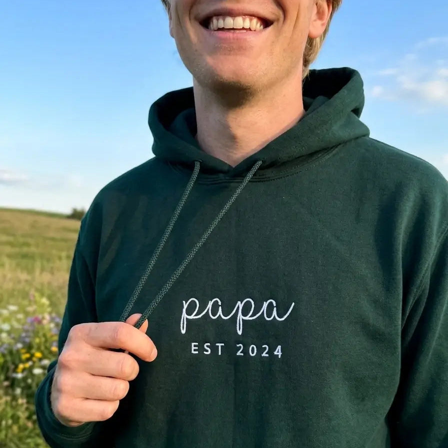 Ein Mann trägt einen personalisierten Hoodie mit einer Stickerei vorne: papa est 2024.