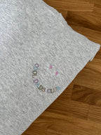 Ein grauer Hoodie liegt auf einem Holztisch. Auf der Brust ist ein lächelndes Gesicht aufgestickt, wobei der Mund aus dem Schriftzug "COOL DADS CLUB" in bunten Farben besteht und zwei Punkte darüber die Augen darstellen.