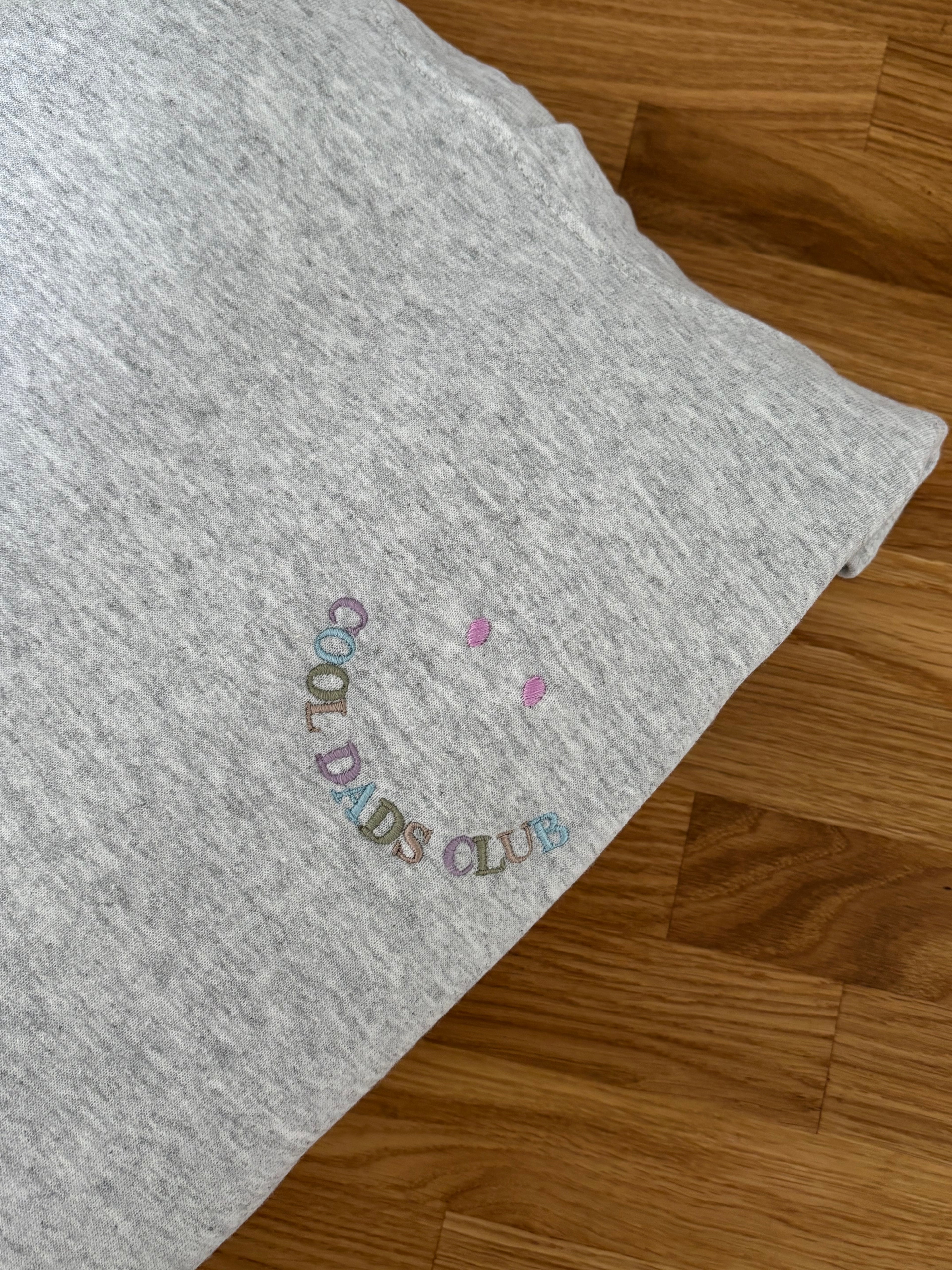 Ein grauer Hoodie liegt auf einem Holztisch. Auf der Brust ist ein lächelndes Gesicht aufgestickt, wobei der Mund aus dem Schriftzug "COOL DADS CLUB" in bunten Farben besteht und zwei Punkte darüber die Augen darstellen.