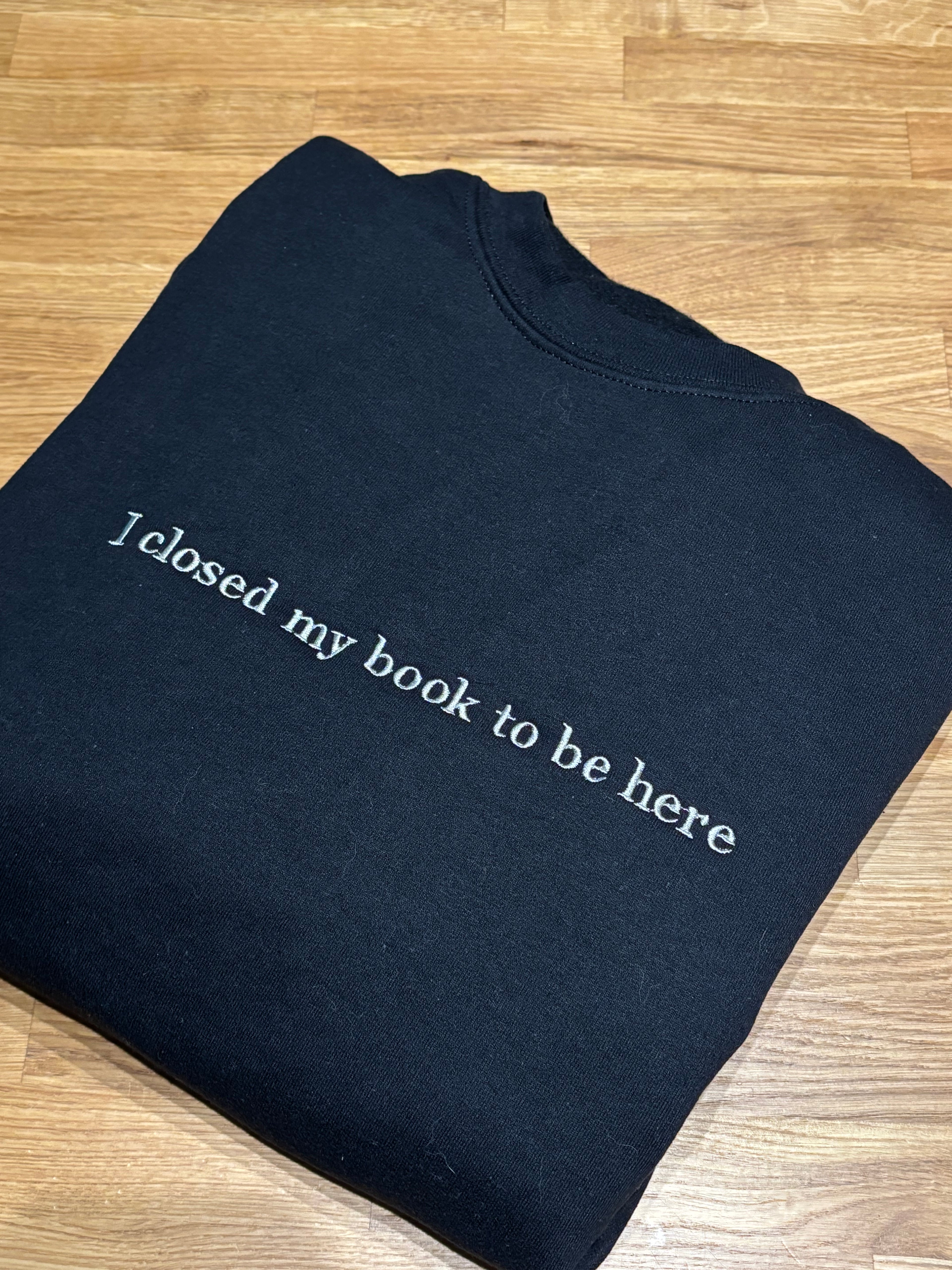 Ein schwarzer Hoodie mit einem grauen Stick auf der Brust "I closed my book to be here".