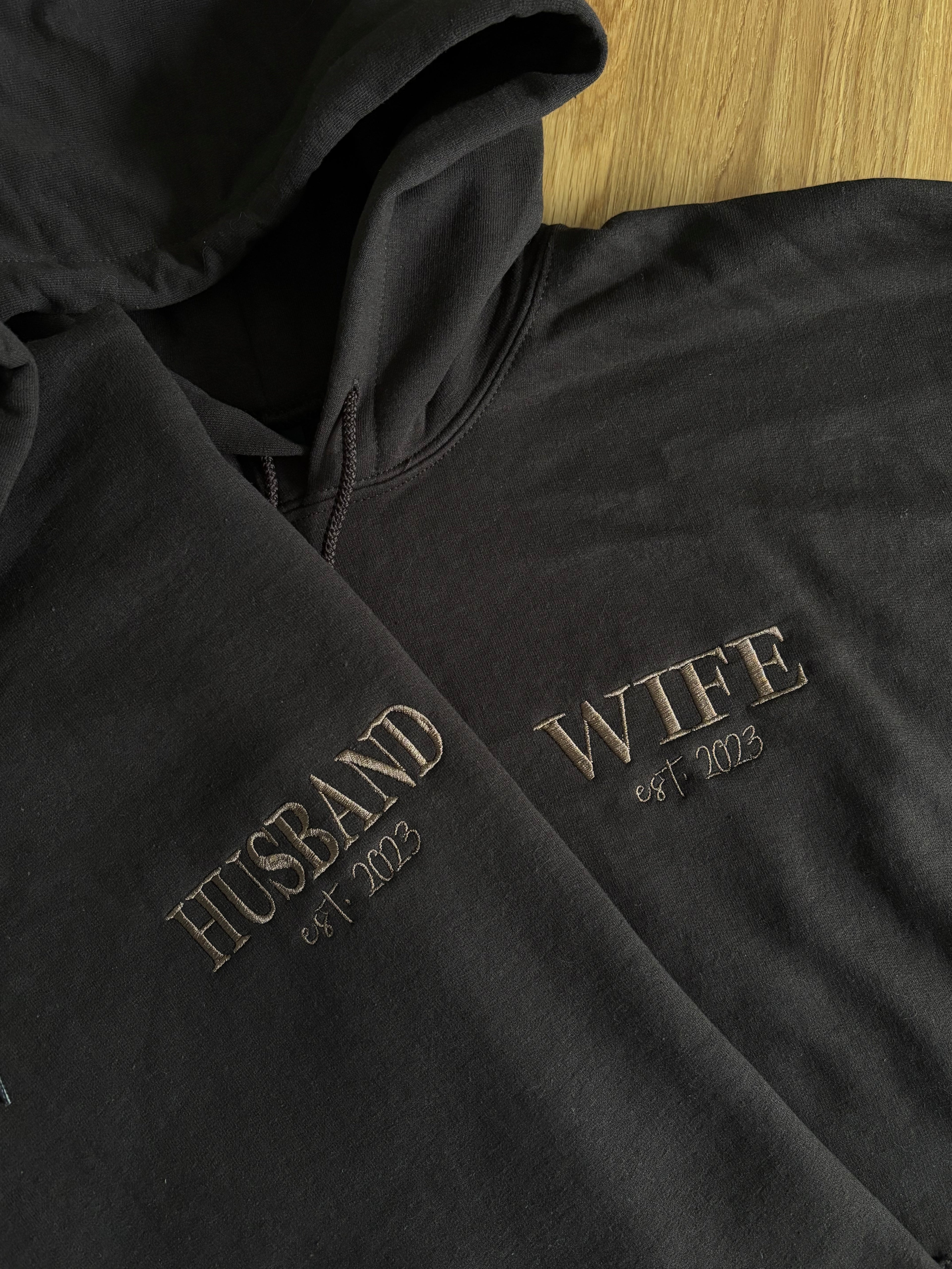 Zwei Pullover mit Kapuze in der Farbe Dark Chocolate. Auf der Brust ist links gestickt "Husband est. 2023" und "Wife est. 2023".