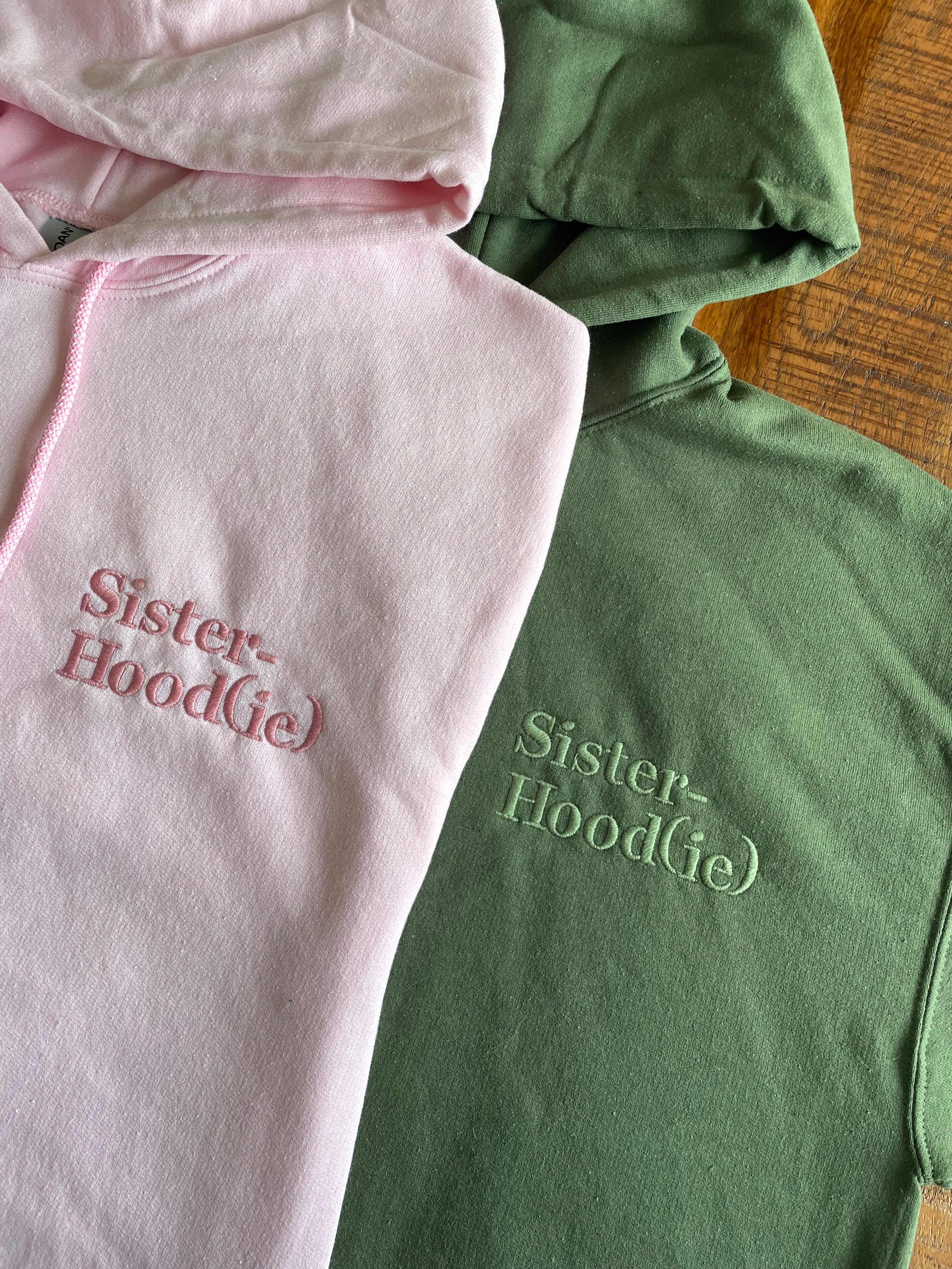 Zwei Pullover mit Kapuze in military green und rosa. Auf der linken Brust steht der Wunschtext "Sister Hood(ie)".