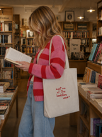 Frau in rosa-rotem Cardigan stöbert mit Jutebeutel in gemütlicher Buchhandlung durch ein Buch.