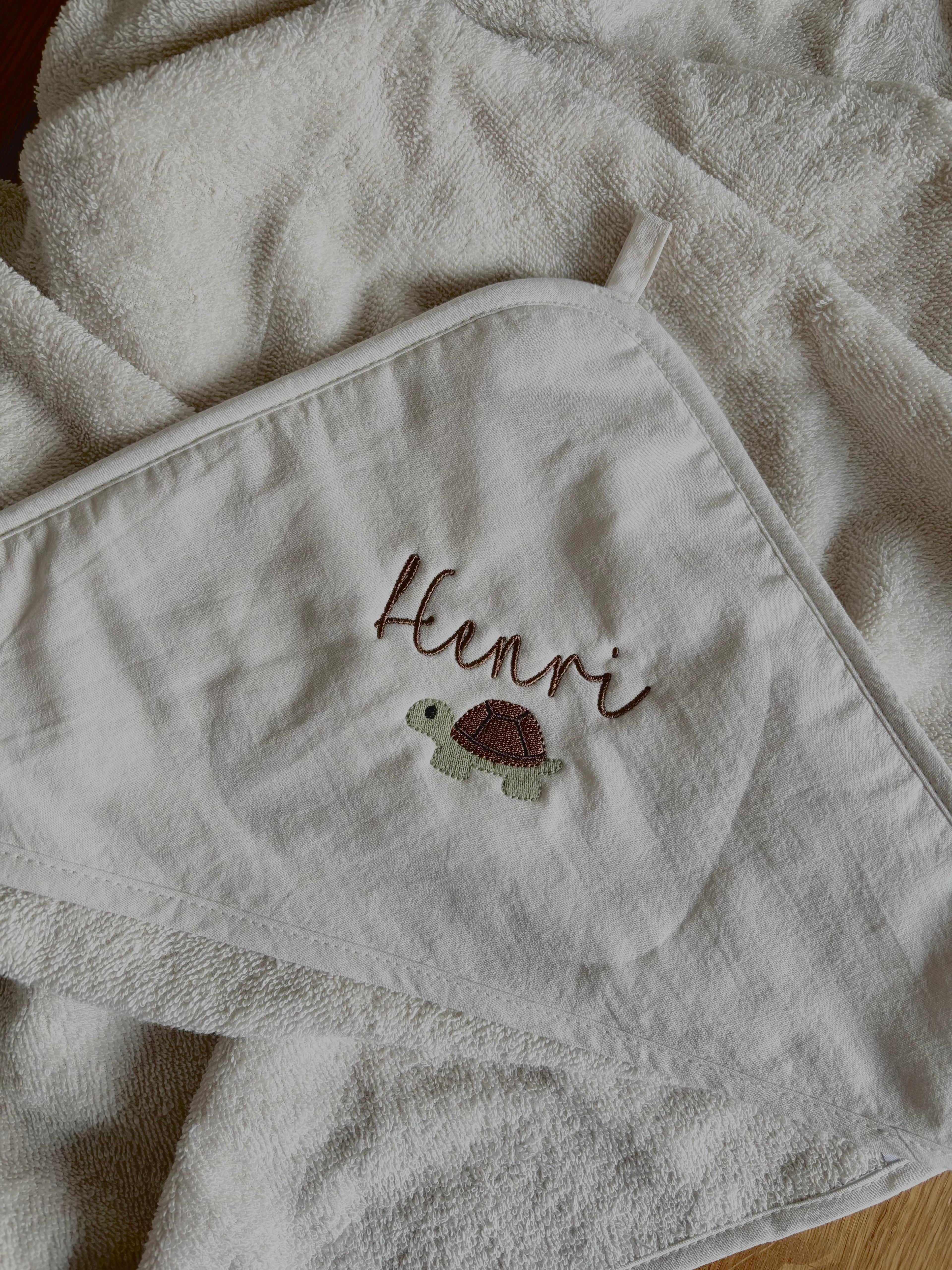 Ein creme farbiges Kapuzenhandtuch mit einem braunen Stick drauf. Auf der Kapuze steht der Name Henri und darunter ist eine Schildkröte in grün/braun.