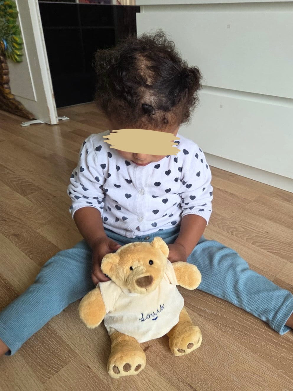 Ein kleines 1-jähriges Kind sitzt auf dem Boden. Vor ihr ist ein bestickter Teddybär, der personalisiert ist. 
