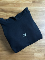 Ein schwarzee Pullover mit Kapuze für Kinder wo die die Initialen "JK" gestickt worden sind.