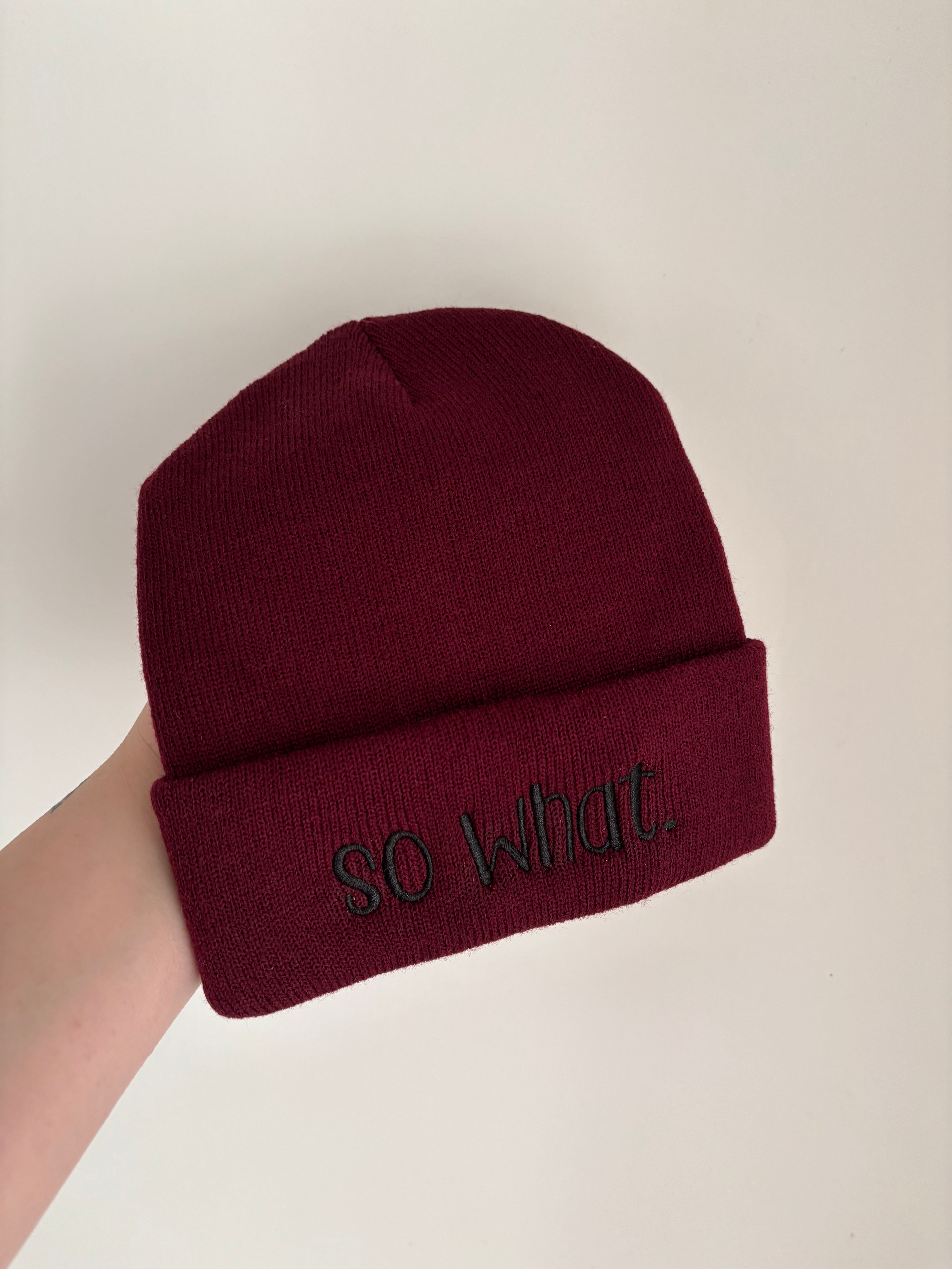 Eine Kindermütze in der Farbe Burgundy mit einer Stickerei vorne in Schwarz: so what.