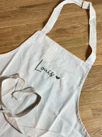 Die Kinderschürze mit einer grünen Stickerei vorne mittig "Louis" mit einem kleinen Herz. 