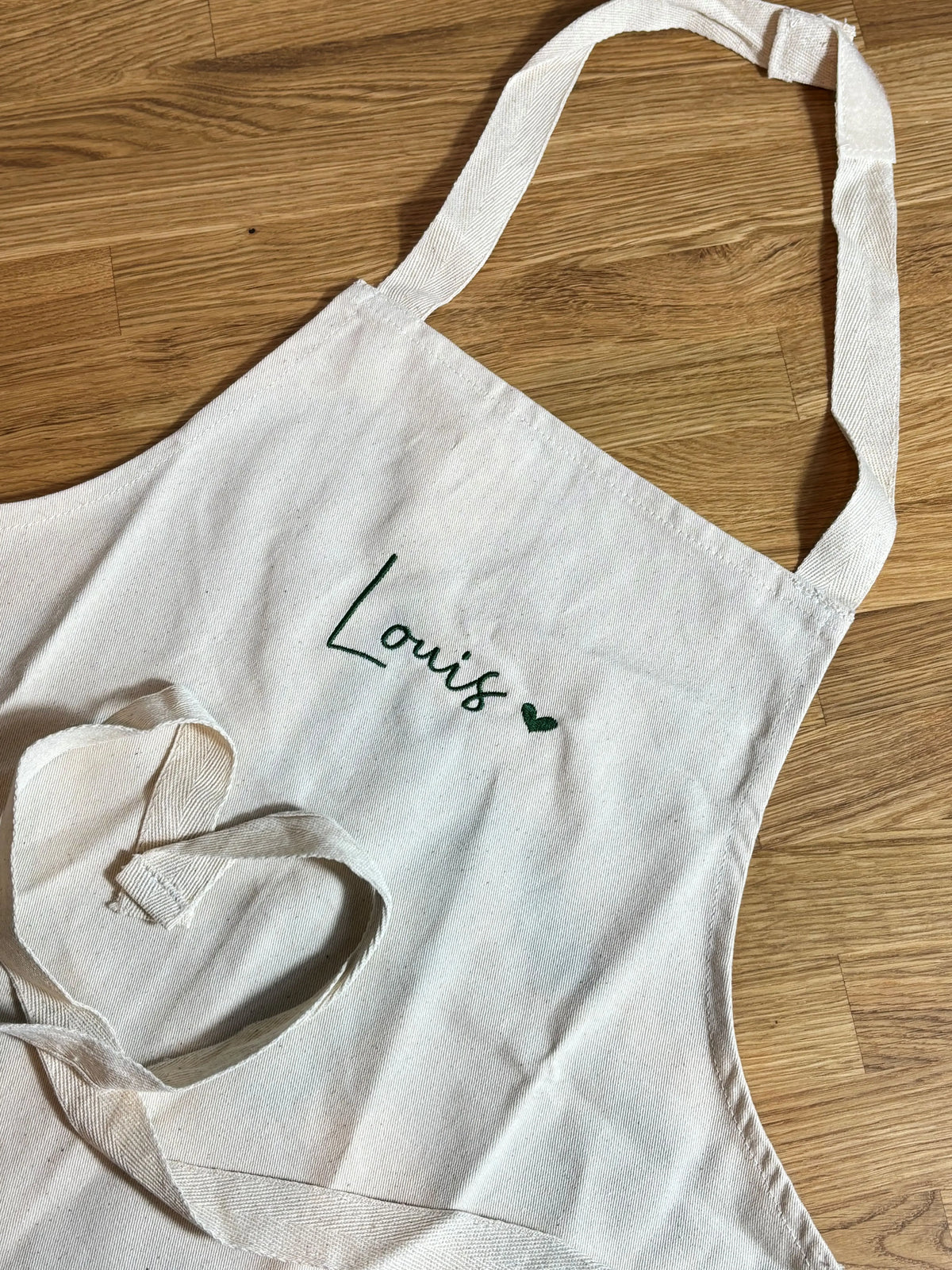Die Kinderschürze mit einer grünen Stickerei vorne mittig "Louis" mit einem kleinen Herz. 