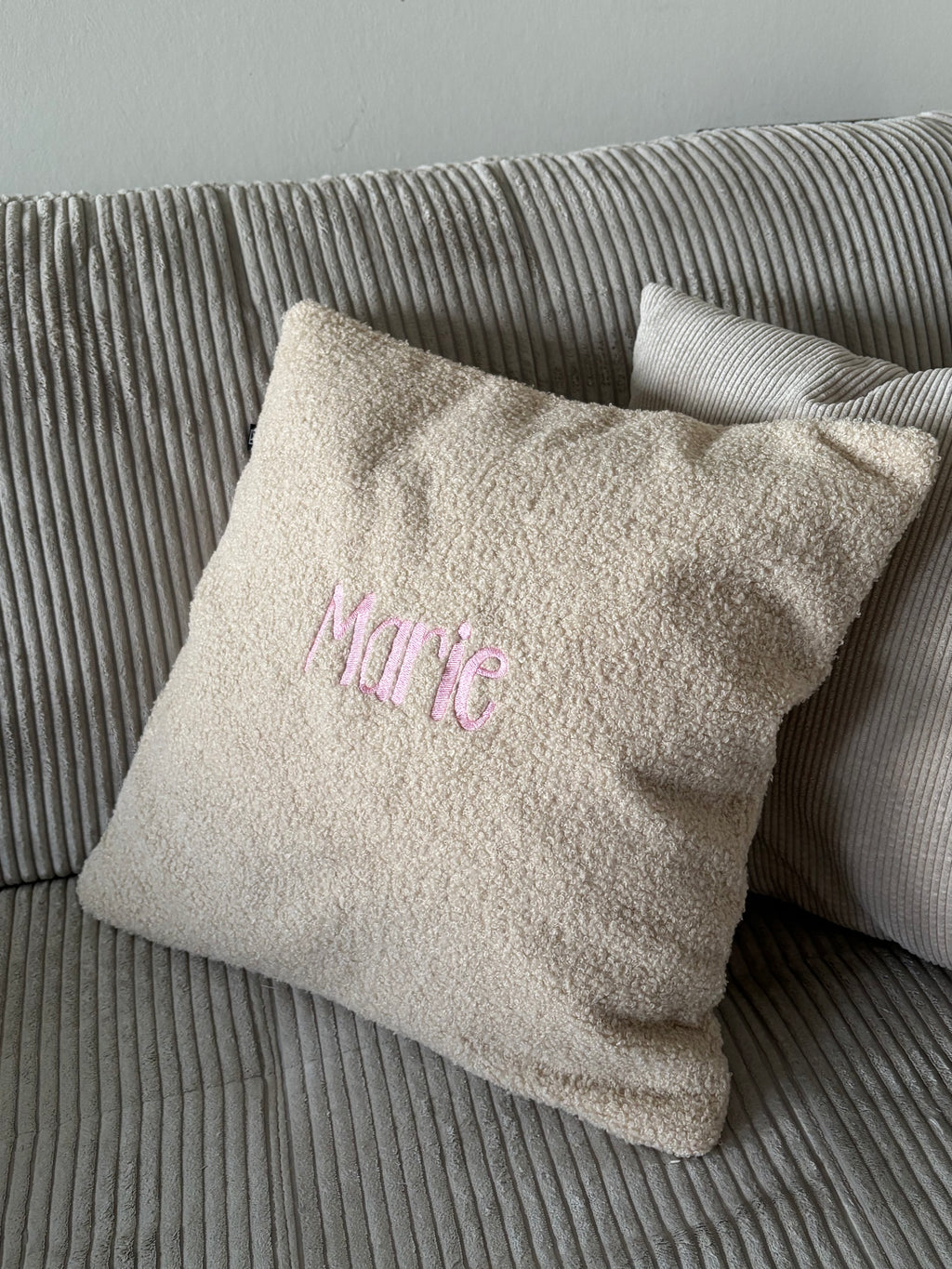 Ein beiges Teddy Kissen auf einem Cord-Sofa. Das perfekte Geschenk für Kinder. Auf der Kissenhülle ist der Name Marie in Rosa gestickt. 