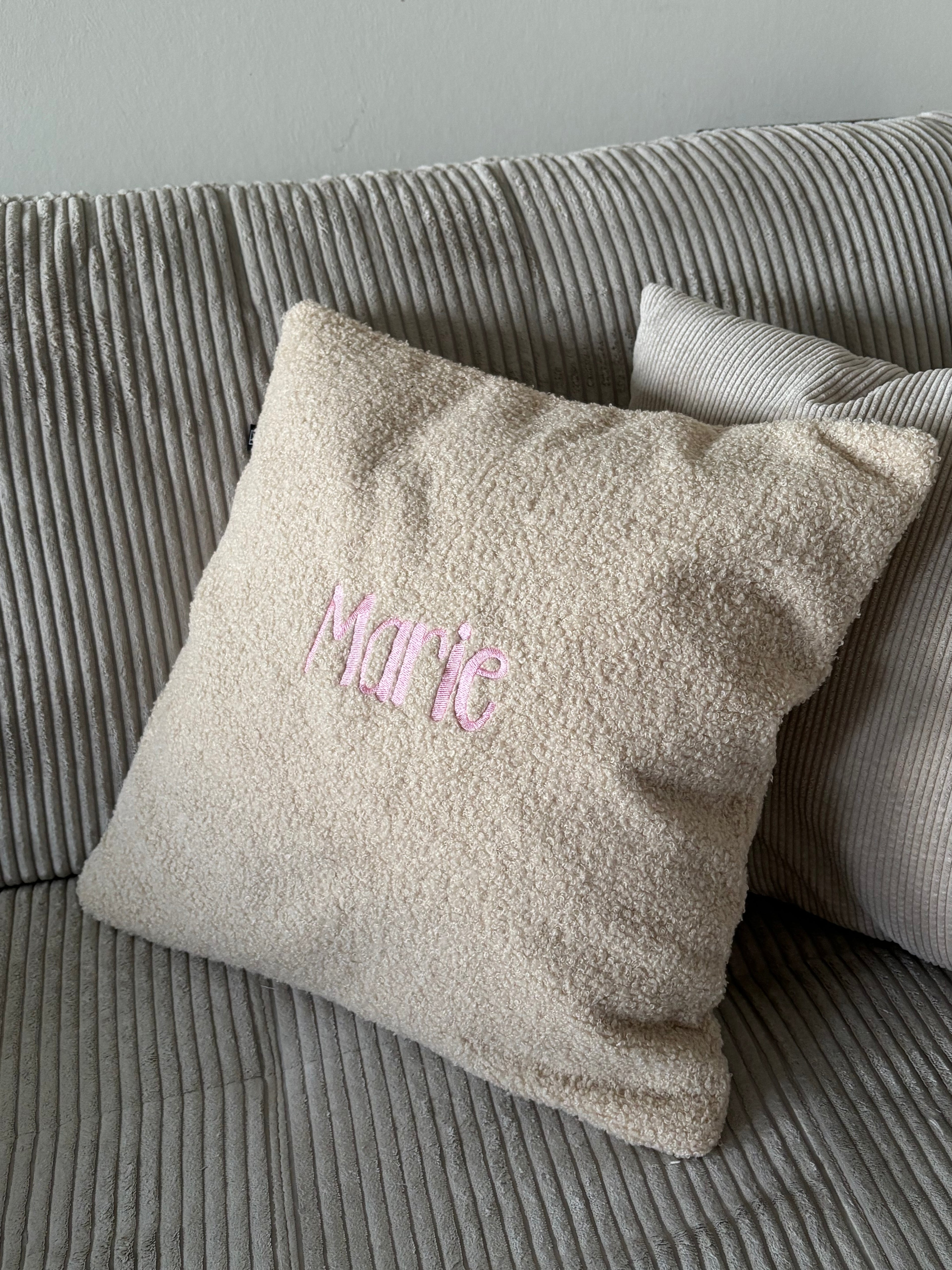 Ein beiges Teddy Kissen auf einem Cord-Sofa. Das perfekte Geschenk für Kinder. Auf der Kissenhülle ist der Name Marie in Rosa gestickt. 