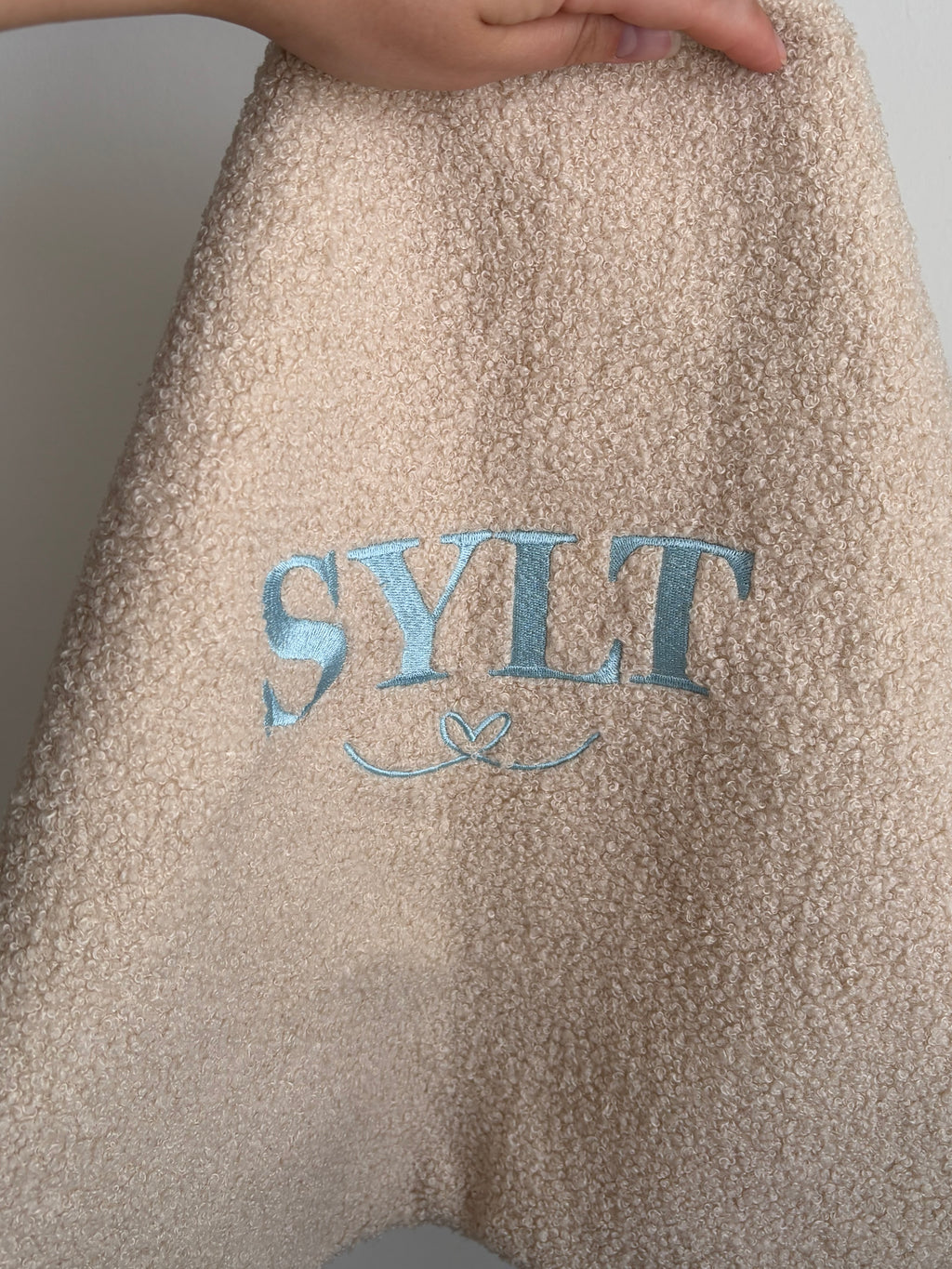 Ein beiges Teddy Kissen mit der Bestickung Sylt in Hellblau. Darunter ist ein verschnörkeltes Herz gestickt. Das perfekte Geschenk für ein schönes Zuhause. 