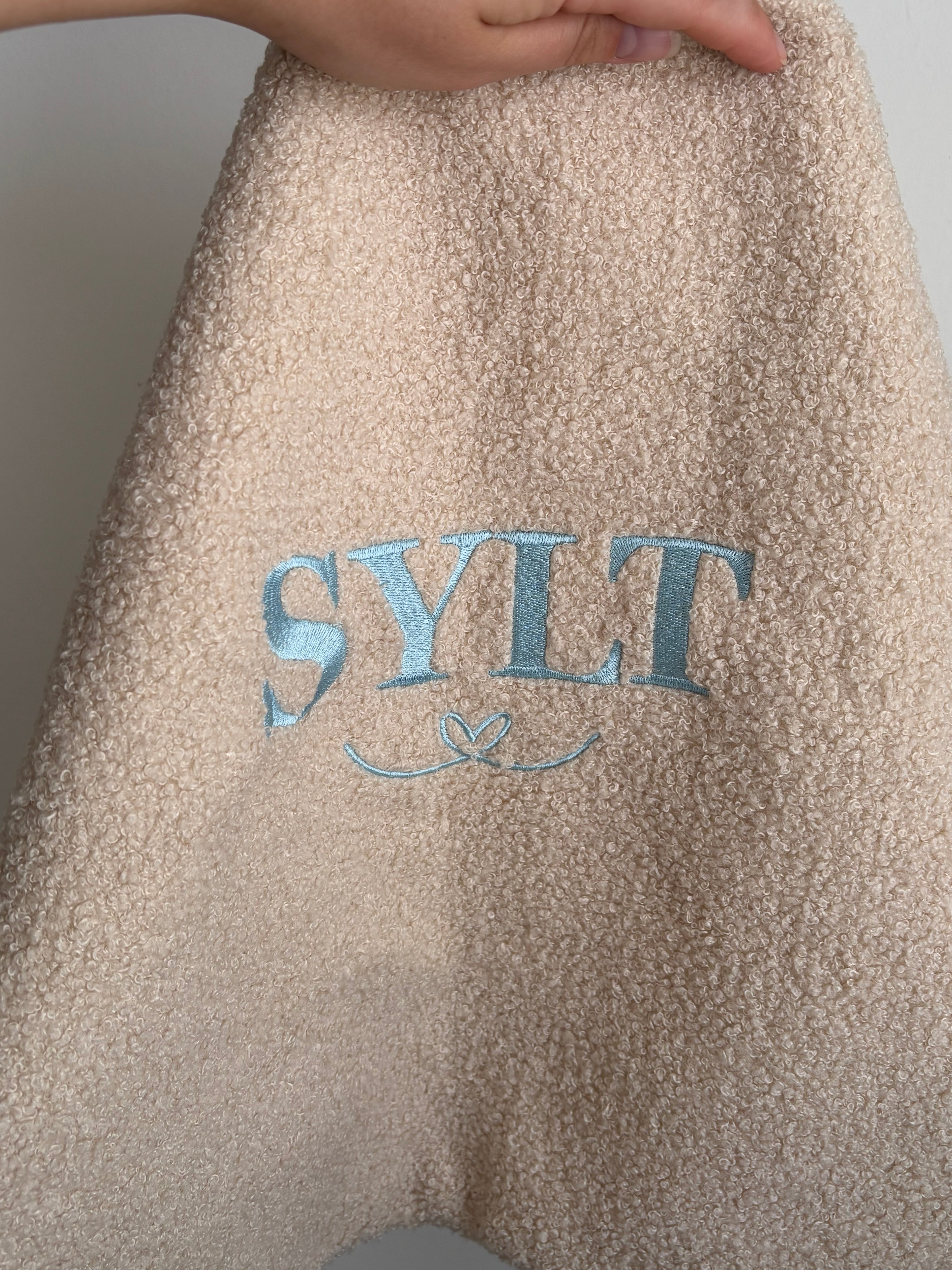 Ein beiges Teddy Kissen mit der Bestickung Sylt in Hellblau. Darunter ist ein verschnörkeltes Herz gestickt. Das perfekte Geschenk für ein schönes Zuhause. 