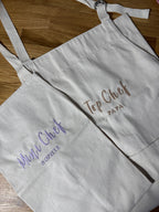 Zwei Schürzen sind zu sehen. Die Schürze für Kinder ist mit Mini Chef Sophie bestickt. Die Schürze für Erwachsene ist mit Top Chef Papa bestickt.