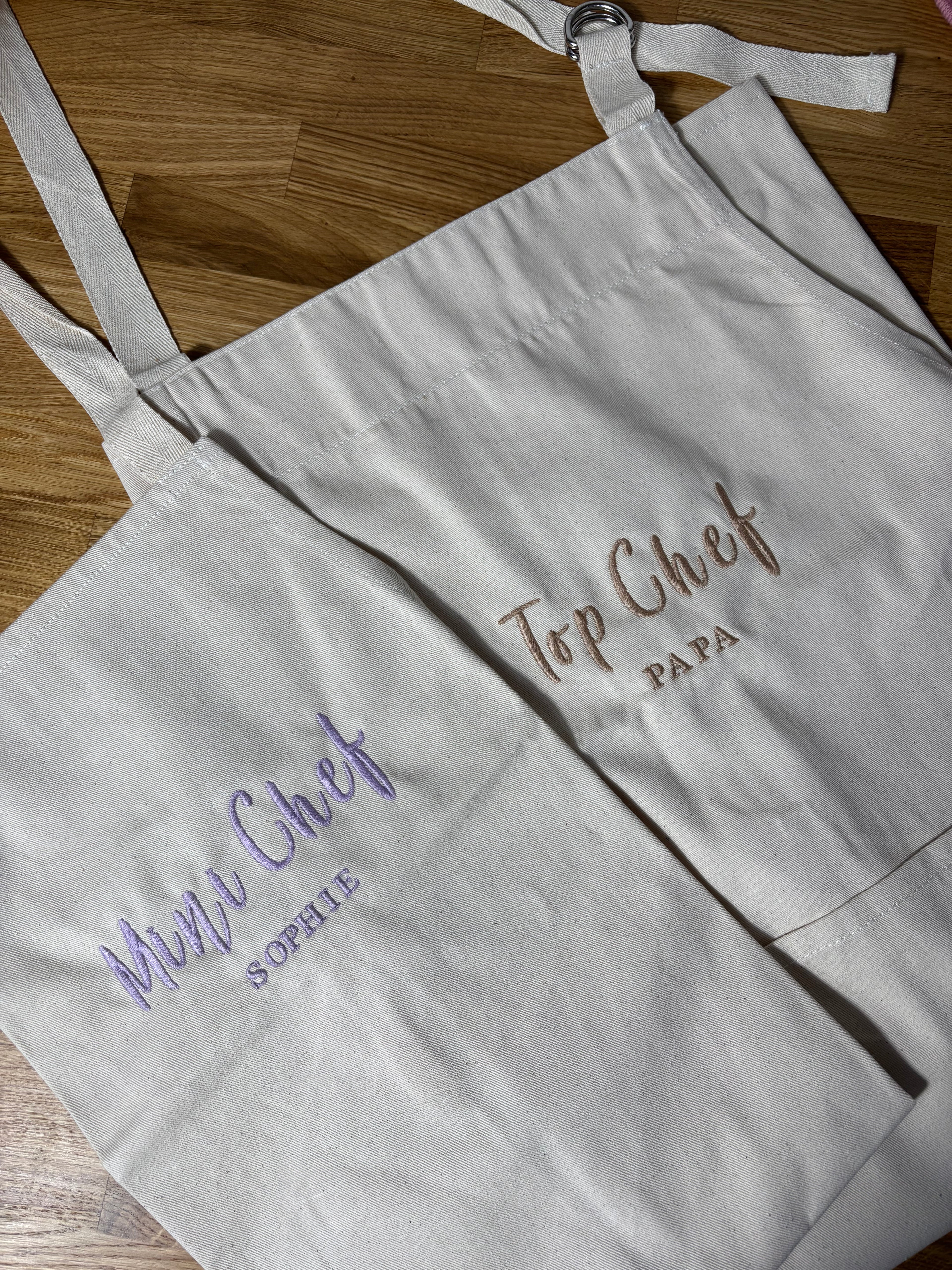 Zwei Schürzen sind zu sehen. Die Schürze für Kinder ist mit Mini Chef Sophie bestickt. Die Schürze für Erwachsene ist mit Top Chef Papa bestickt.