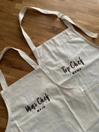 Zwei Schürzen jeweils mit braunem Garn bestickt. Die Junior Schürze ist mit Mini Chef Maja bestickt und die Erwachsenen Schürze mit Top Chef Mama.