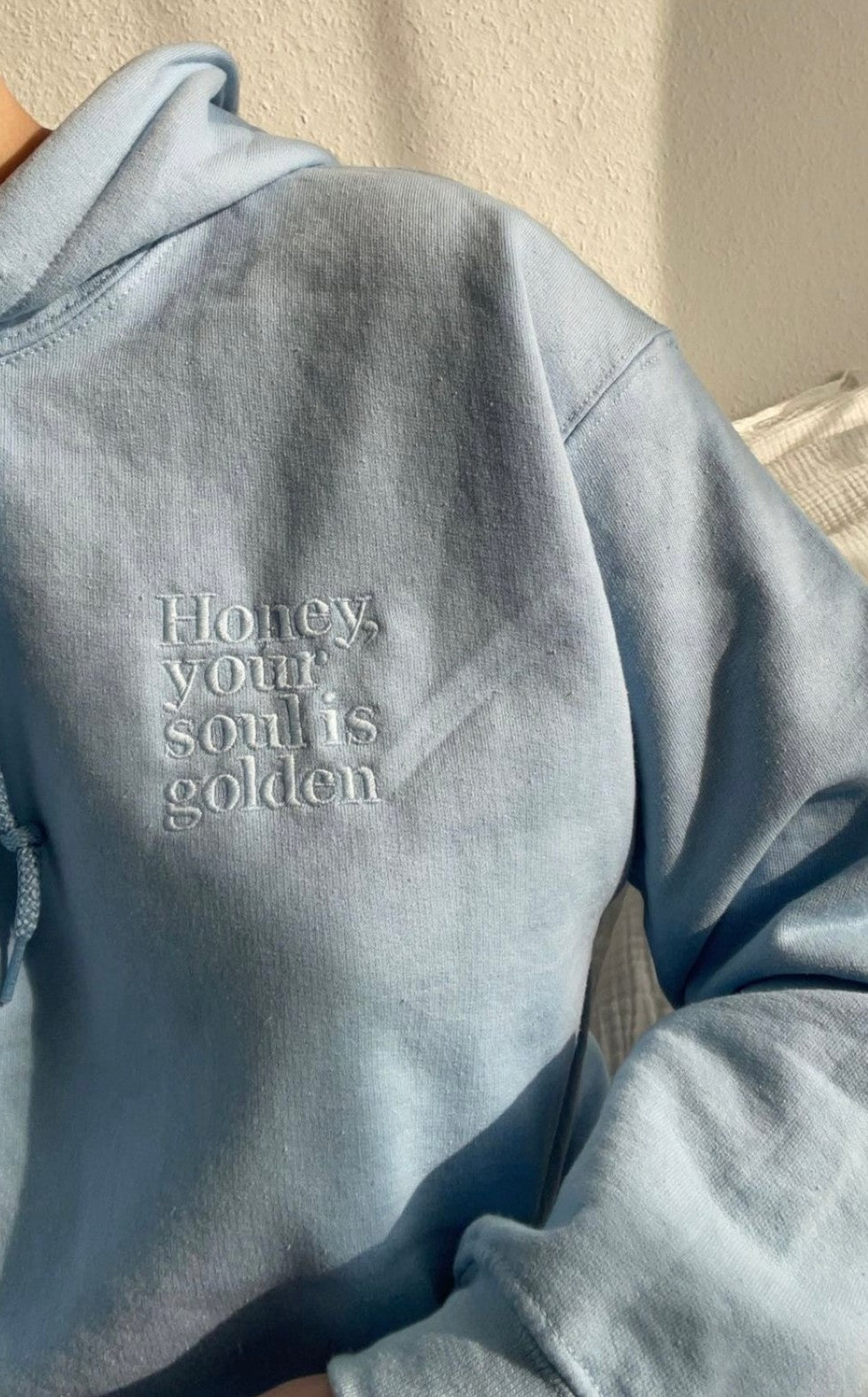Ein Pullover mit Kapuze in der Farbe Light Blue. Vorne links auf der Brust ist der Pullover bestickt mit "Honey, your soul is golden."
