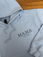 Ein Hoodie in der Farbe Light Blue mit einer Stickerei in der Farbe Stone Blue: Mama est. 2026.