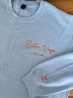 Ein Sweater in der Farbe Light Blue mit einer Stickerei in der Farbe Orange: Bester Papa est. 2025. Auf dem linken Ärmel sind zwei Kindernamen gestickt: Elli und Lou.