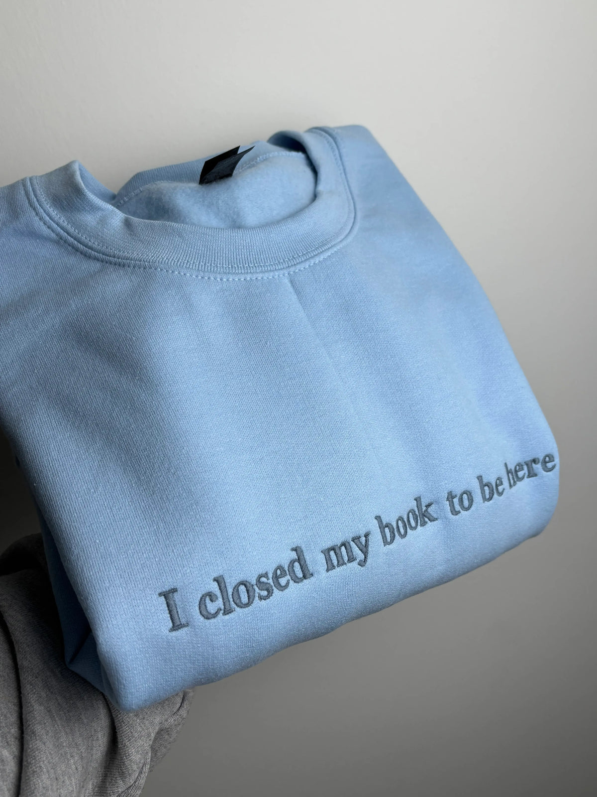 Ein Sweater in der Farbe Light Blue mit dem Schriftzug I closed my Book To be Here. Der Sweater ist mit einem dunkelblauen Garn bestickt.