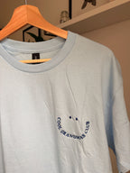 Ein T-Shirt in der Farbe Light Blue mit kurzen Ärmeln und einen dunkelblauen Smiley auf der linken Brustseite. Die Grafik besteht aus zwei kleinen ovalen Augen und einem geschwungenen Schriftzug COOL GRANDMAS CLUB, der ein lächelndes Gesicht bildet.
