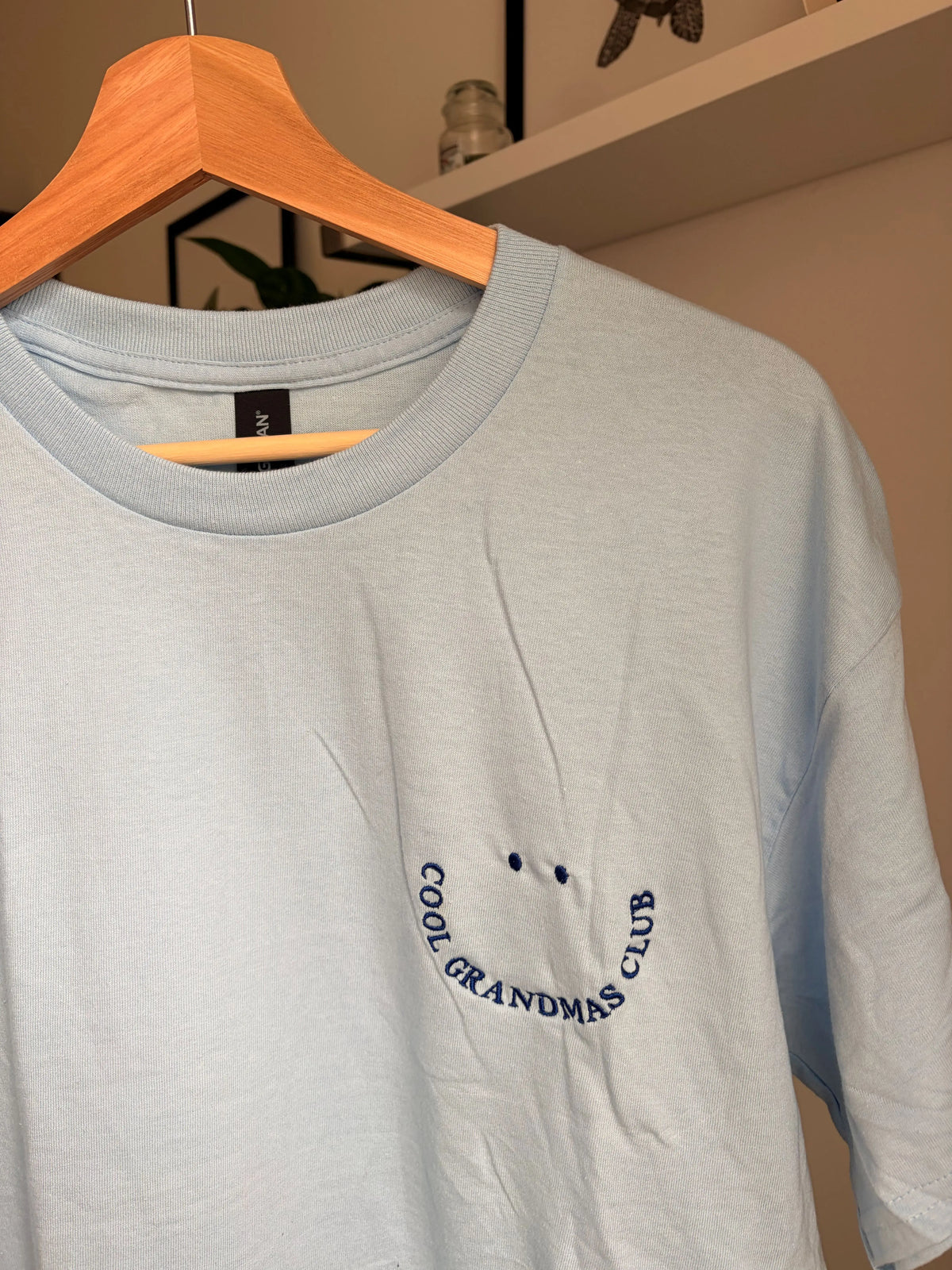 Ein T-Shirt in der Farbe Light Blue mit kurzen Ärmeln und einen dunkelblauen Smiley auf der linken Brustseite. Die Grafik besteht aus zwei kleinen ovalen Augen und einem geschwungenen Schriftzug COOL GRANDMAS CLUB, der ein lächelndes Gesicht bildet.