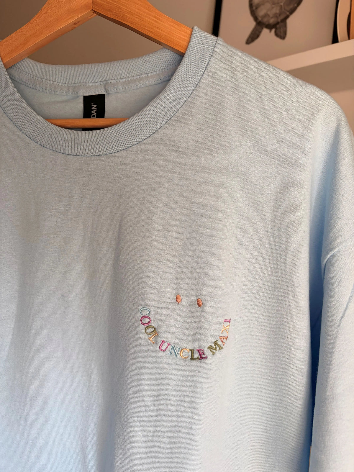 Ein hellblaues T-Shirt mit Rundhalsausschnitt und einer Stickerei auf der linken Brust. Das Design besteht aus zwei kleinen lila Punkten als Augen über einer geschwungenen Linie aus mehrfarbigem Text, der COOL UNCLE MAXI bildet.