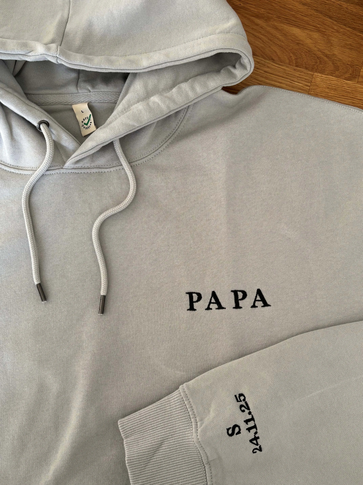 Der Pullover in der Farbe Light Grey ist mit einem schwarzen Garn bestickt: Papa. Auf dem linken Ärmel ist in schwarz eingestickt: S 24.11.25.