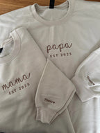 Zwei beige Sweater für Eltern. Beide sind jeweils mit Mama und Papa bestickt worden. Auf dem Ärmel tragen sie beide den Kindernamen Thomas.