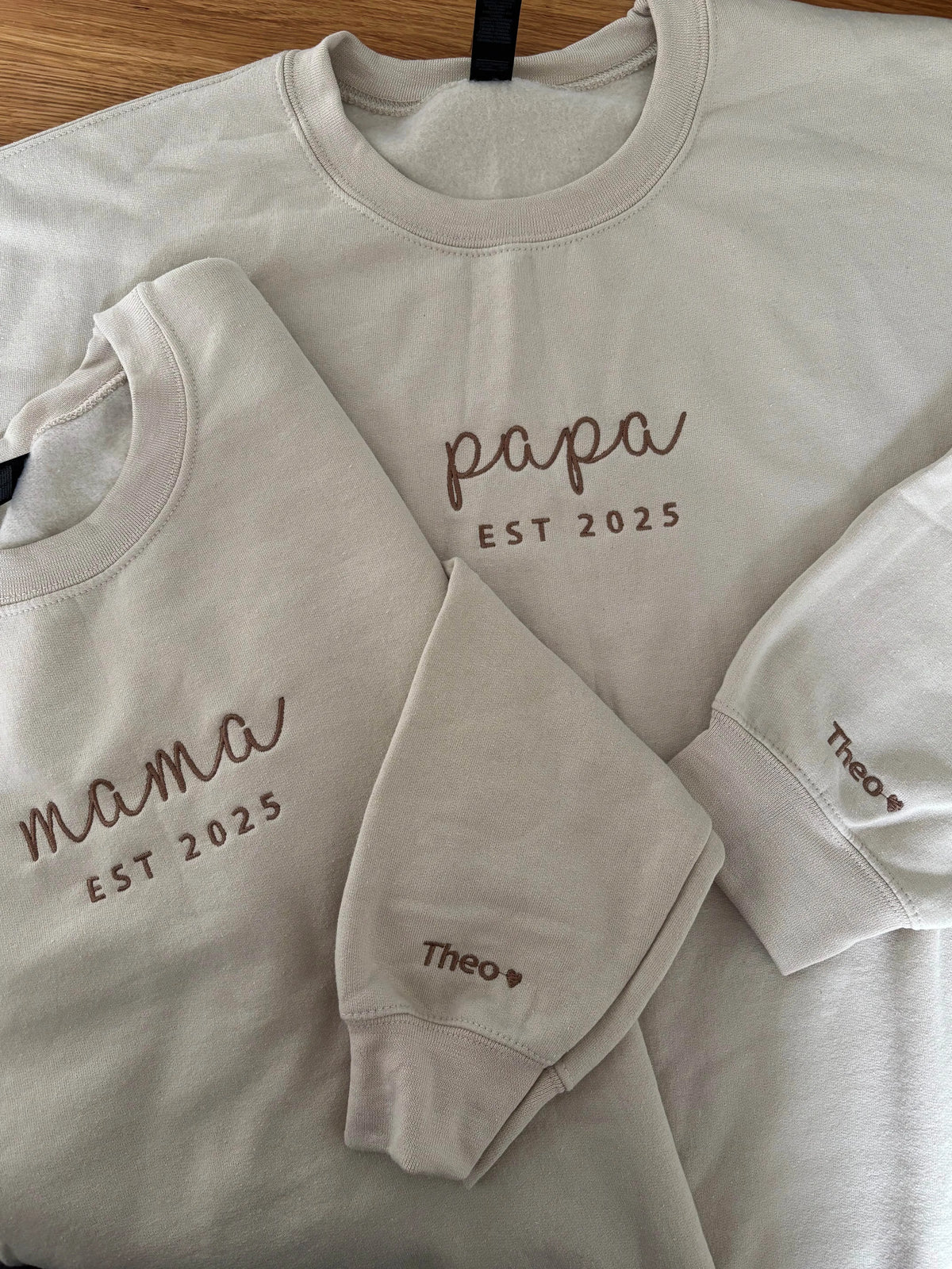 Zwei beige Sweater für Eltern. Beide sind jeweils mit Mama und Papa bestickt worden. Auf dem Ärmel tragen sie beide den Kindernamen Thomas.
