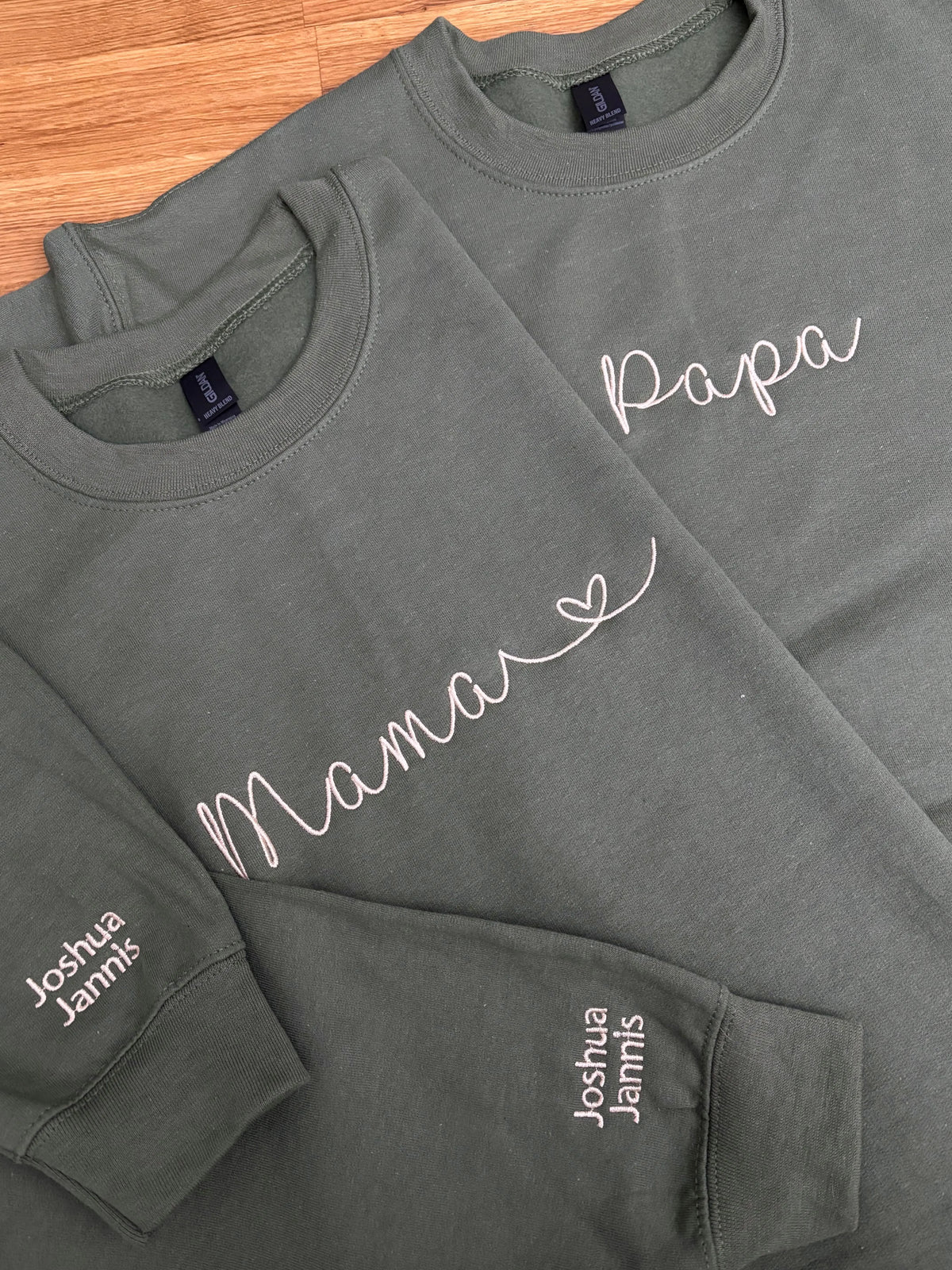 Zwei Sweater in der Farbe Military Greenfür Eltern. Beide sind jeweils mit Mama und Papa bestickt worden. Auf dem Ärmel tragen sie beide die Kindernamen Joshua und Jannis.