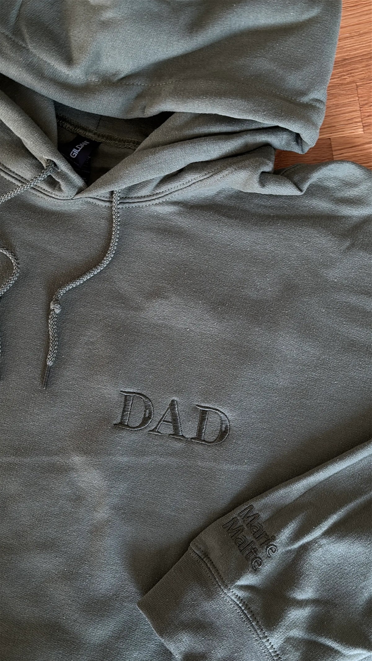 Ein Hoodie in der Farbe Military Green mit einer Stickerei in der Farbe Green: Dad. Auf dem linken Ärmel sind zwei Kindernamen gestickt: Merle und Malte.
