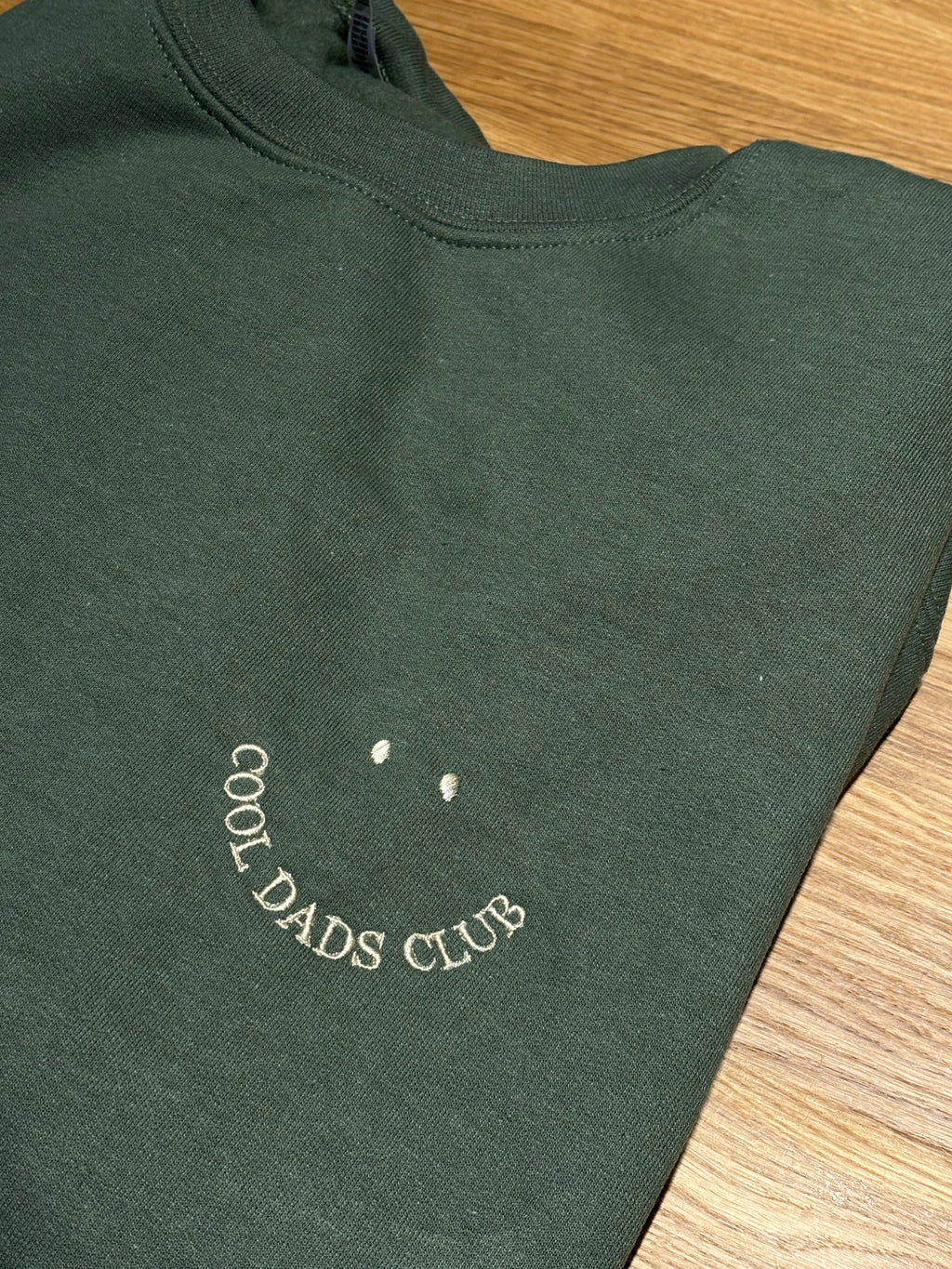 Ein personalisierter Sweater in der Farbe Military Green. Der Stick ist auf der linken Brustseite in beige "Cool Dads Club" als Smiley.