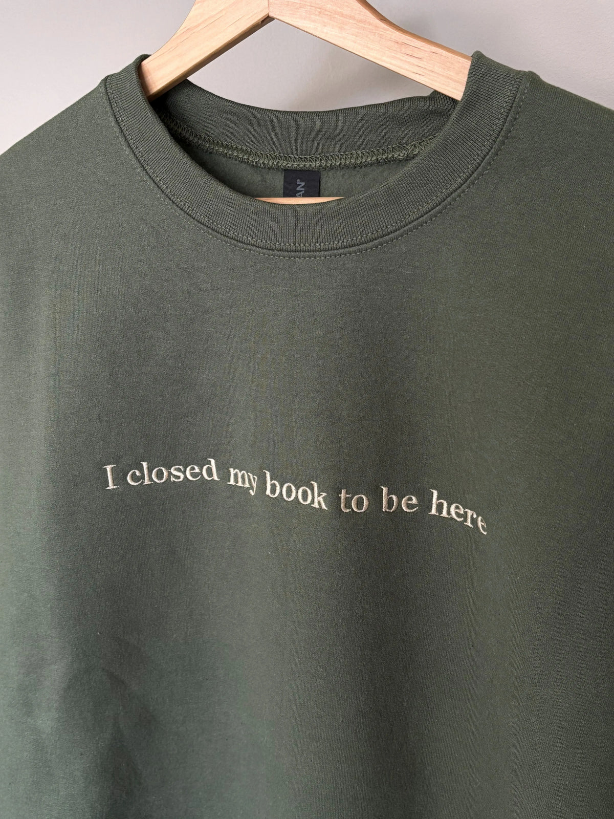 Ein Sweater in der Farbe Military Green mit einer beigen Stickerei vorne mittig auf der Brust: I closed my book to be here.