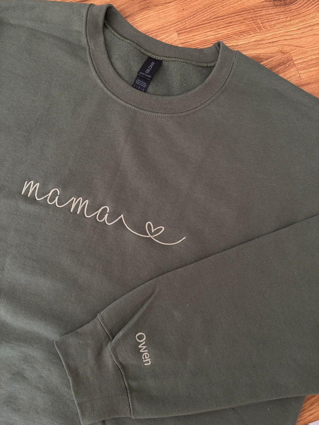 Ein personalisierter Sweater in der Farbe military green. Auf der Brust ist in moss "mama" mit Herz gestickt. Auf dem Ärmel ist ein individueller Name "Owen" gestickt.