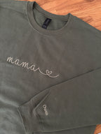 Ein personalisierter Sweater in der Farbe military green. Auf der Brust ist in moss "mama" mit Herz gestickt. Auf dem Ärmel ist ein individueller Name "Owen" gestickt.