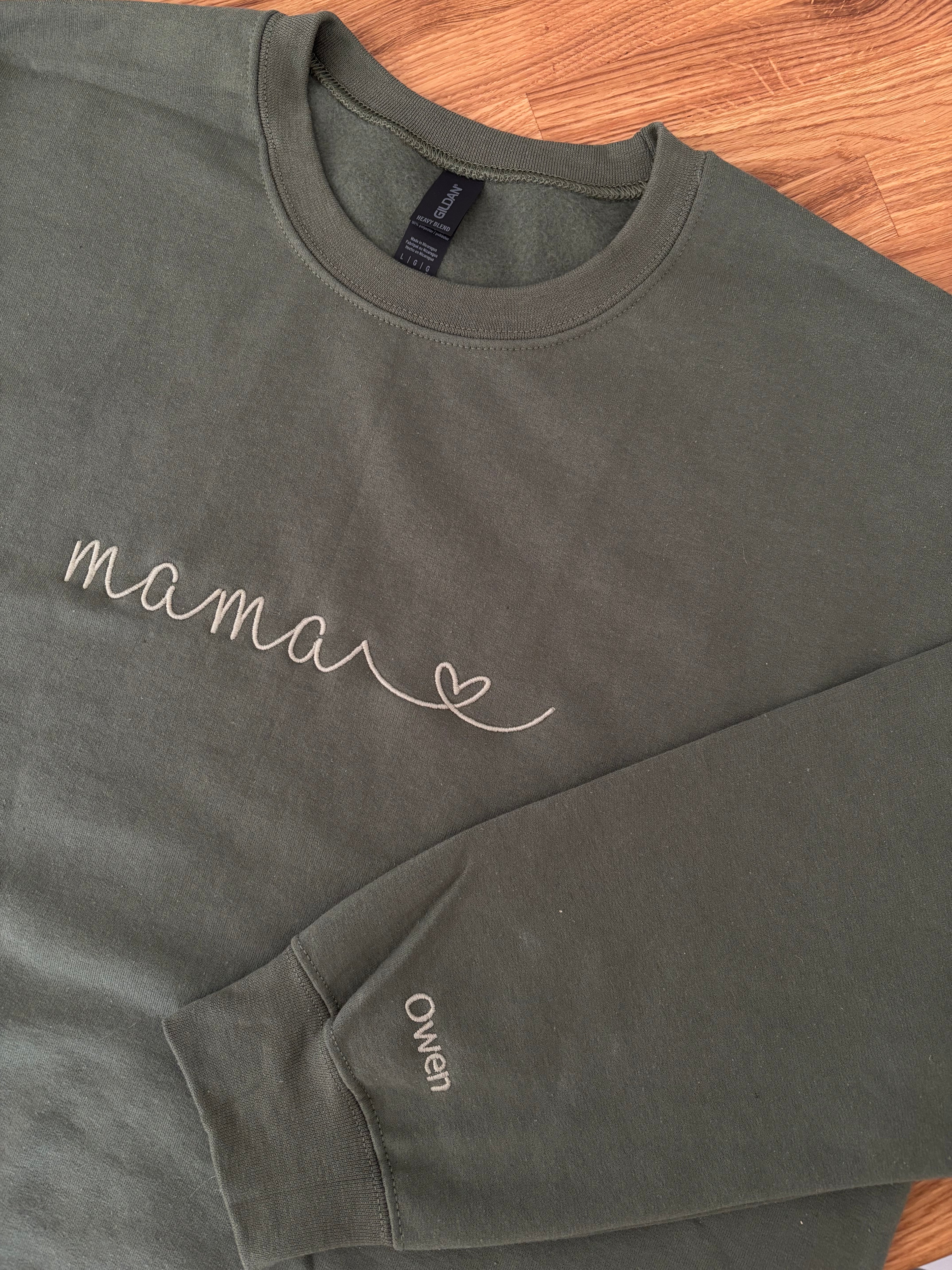 Ein personalisierter Sweater in der Farbe military green. Auf der Brust ist in moss "mama" mit Herz gestickt. Auf dem Ärmel ist ein individueller Name "Owen" gestickt.