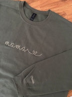 Ein personalisierter Sweater in der Farbe military green. Auf der Brust ist in moss "mama" mit Herz gestickt. Auf dem Ärmel ist ein individueller Name "Owen" gestickt.