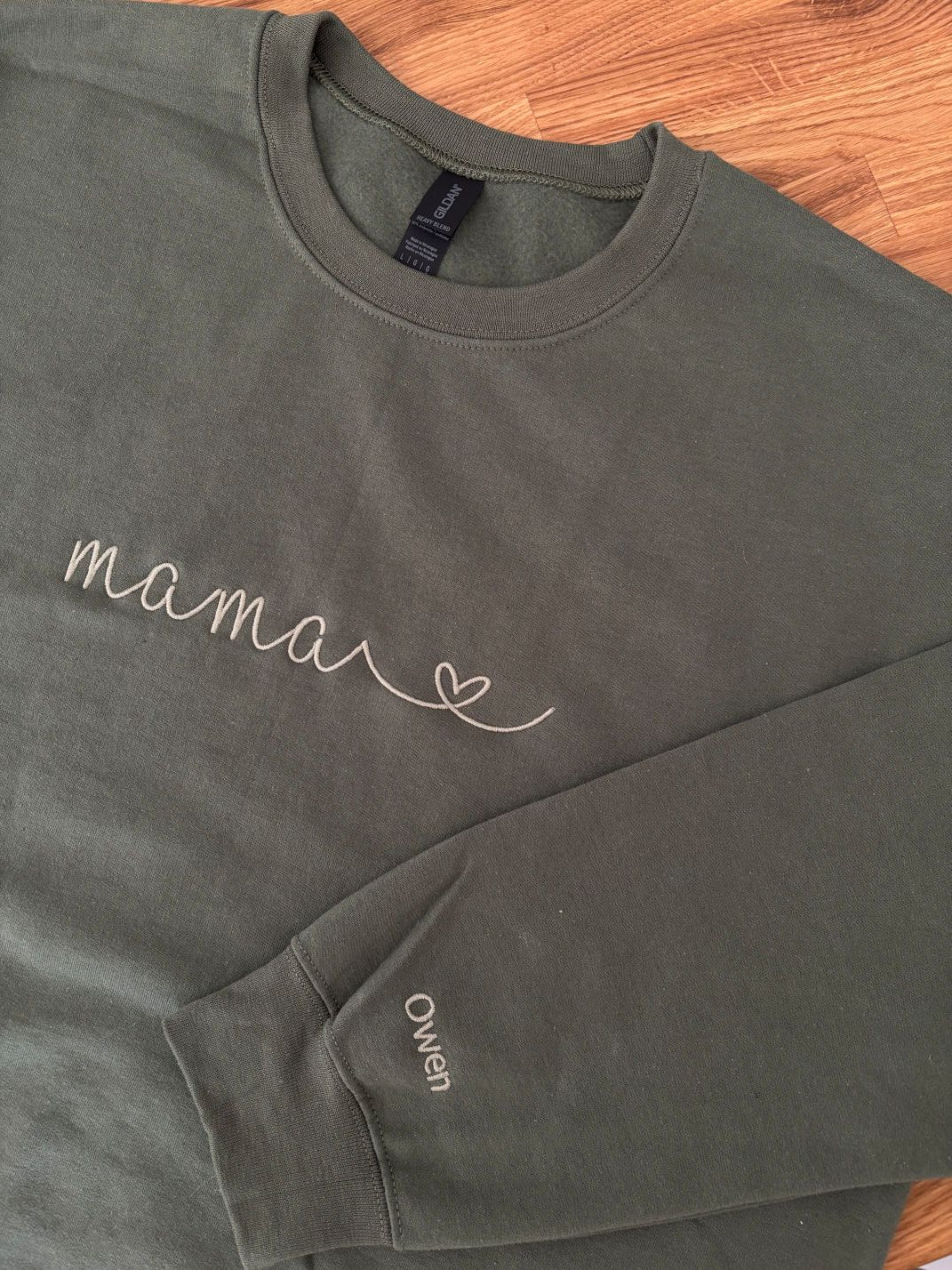Ein personalisierter Sweater in der Farbe military green. Auf der Brust ist in moss "mama" mit Herz gestickt. Auf dem Ärmel ist ein individueller Name "Owen" gestickt.