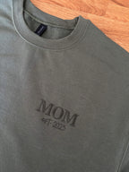 Ein Sweater in der Farbe Military Green mit einer Stickerei in der Farbe Green: MOM est. 2025.