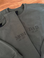 Zwei Sweater in der Farbe Military Green mit jeweils einer Stickerei in der Farbe Green: MOM & DAD est. 2025.