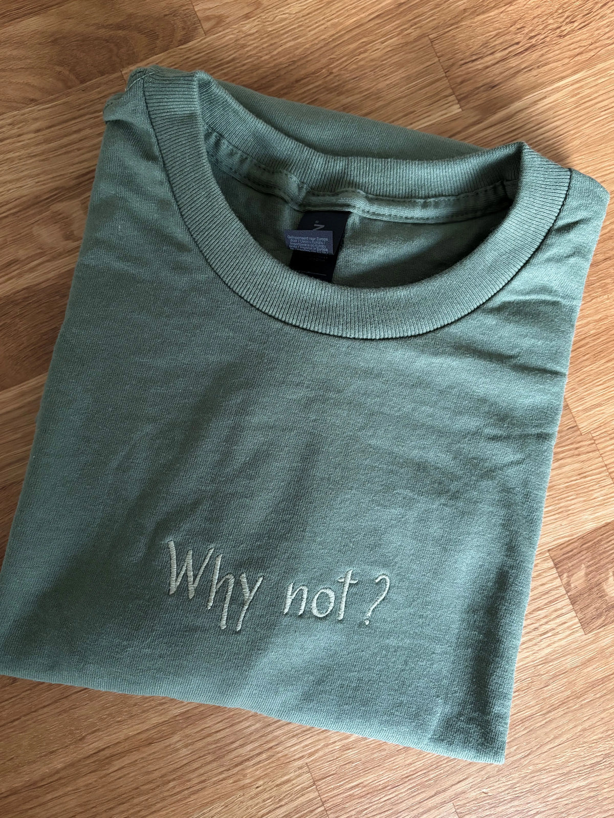 Ein T-Shirt in der Farbe Military Green mit einer Stickerei in der Farbe Moss vorne mittig auf der Brust "Why not?".