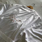 Ein weißer Kimono mit einem Gürtel. Auf dem Rücken ist Braut Name gestickt. Der Mantel liegt auf einem Bett.