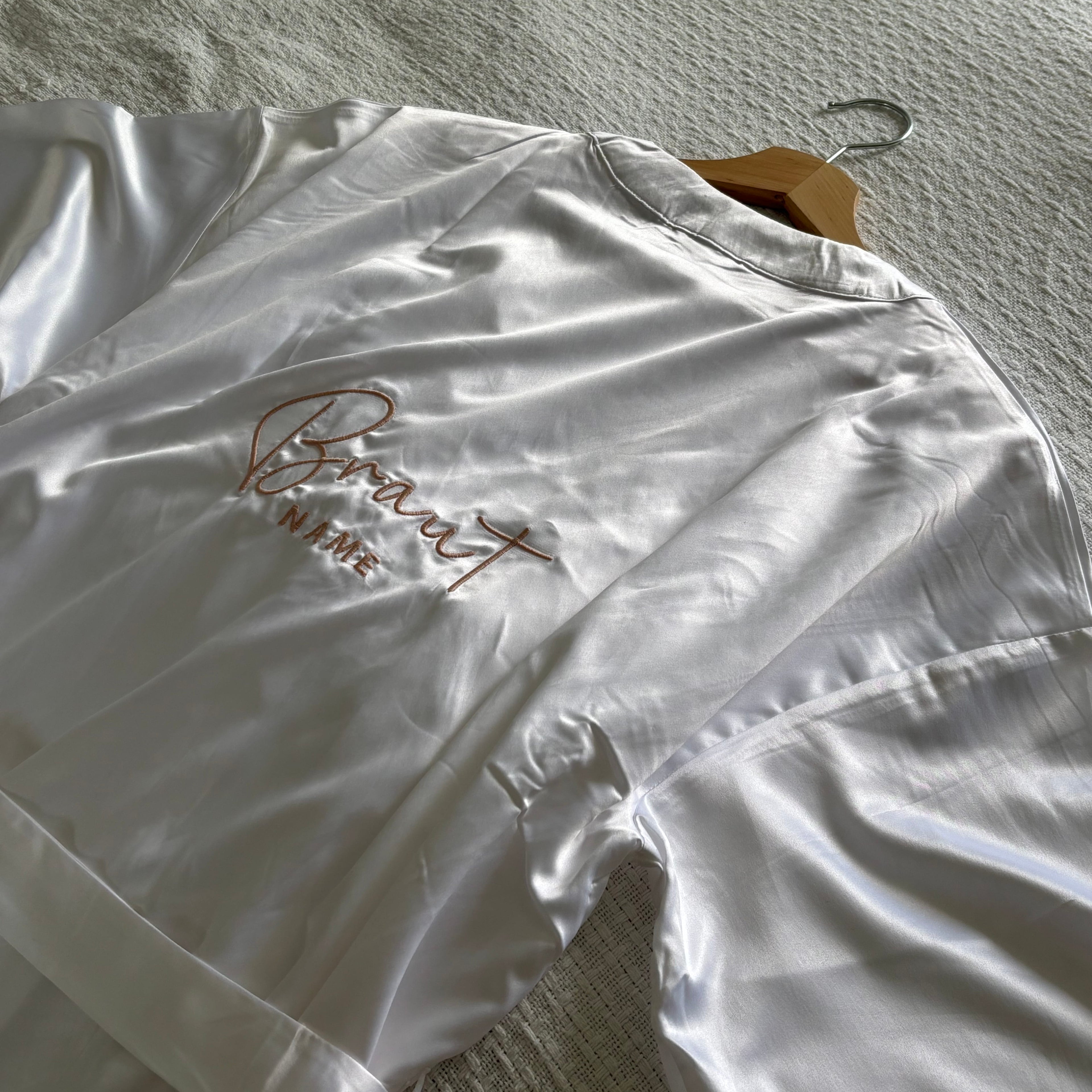 Ein weißer Kimono mit einem Gürtel. Auf dem Rücken ist Braut Name gestickt. Der Mantel liegt auf einem Bett.