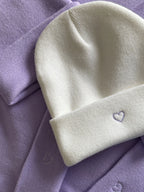Es liegen mehrere Mützen in der Farbe Lavender auf dem Tisch, die mit einem weißen Herz bestickt ist. Außerdem liegt noch eine weiße Mütze mit deinem lavender Herz auf den anderen Mützen drauf.