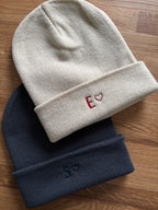 Zwei Mützen in almond und dunkegrau. Vorne ist jeweils ein Anfangsbuchstabe mit einem Herz gestickt.