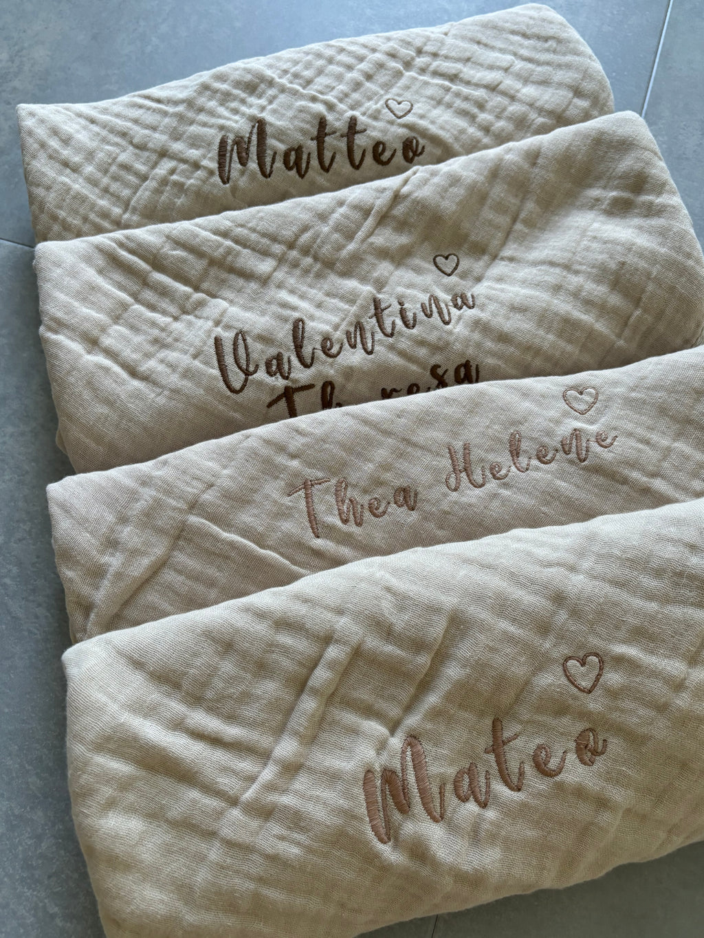 Es sind beige Musselin Spucktücher abgebildet. Auf jedem Tuch ist eine Namensstickerei, welche individuell und personalisiert abgestimmt ist. Zusätzlich zum Namen ist ein Herz gestickt.